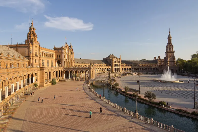 The curved brick and tile facade of the Plaza de España in Seville reflected in its canal